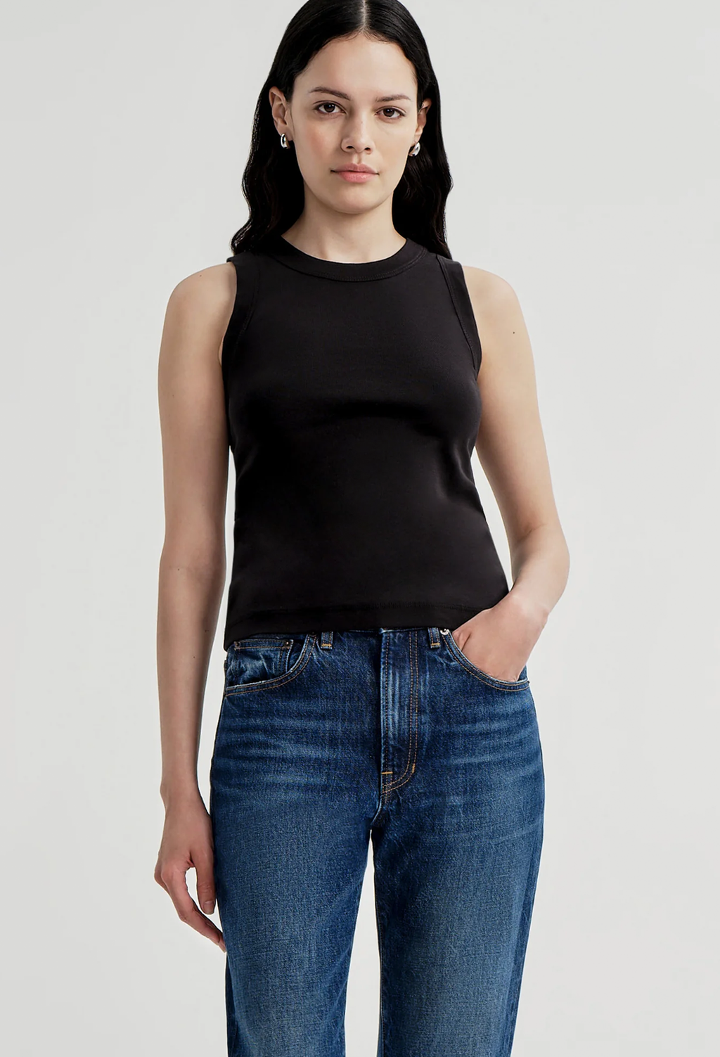 A woman modeling a black sleeveless ribbed knit top paired with blue jeans, photographed against a light gray studio background.