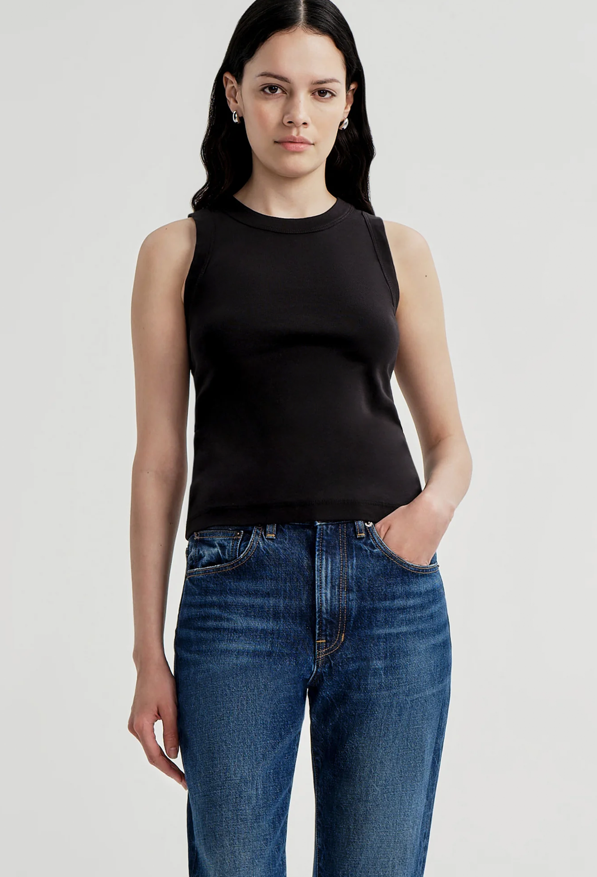 A woman modeling a black sleeveless ribbed knit top paired with blue jeans, photographed against a light gray studio background.