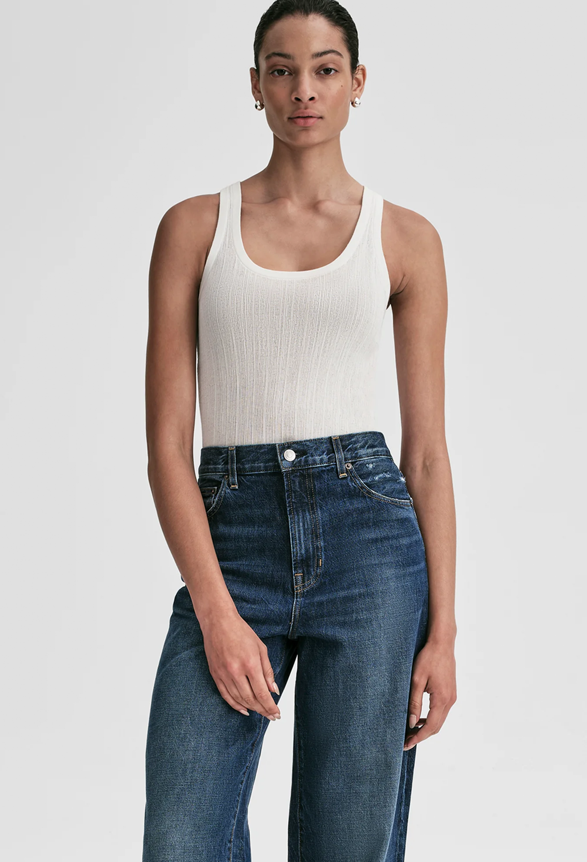 A model wearing a white ribbed knit scoop-neck tank top and dark blue high-waisted jeans, photographed against a solid light gray studio background.