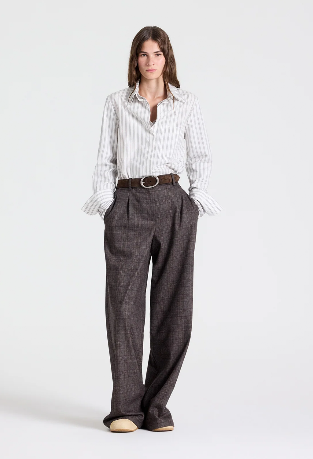 A woman modeling a white and gray vertically striped button-down shirt paired with dark brown wide-leg trousers, a brown belt, and beige shoes, shot against a plain light gray studio background.