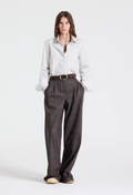 A woman modeling a white and gray vertically striped button-down shirt paired with dark brown wide-leg trousers, a brown belt, and beige shoes, shot against a plain light gray studio background.