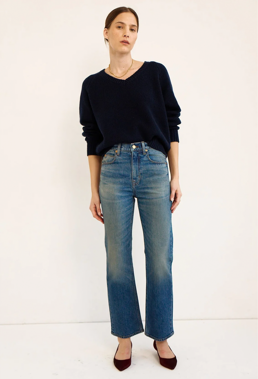 A woman modeling a dark navy blue long-sleeve knit sweater and high-waisted blue jeans with a subtle gradient fade at the hem, paired with dark red pointed-toe heels, against a plain off-white studio background.