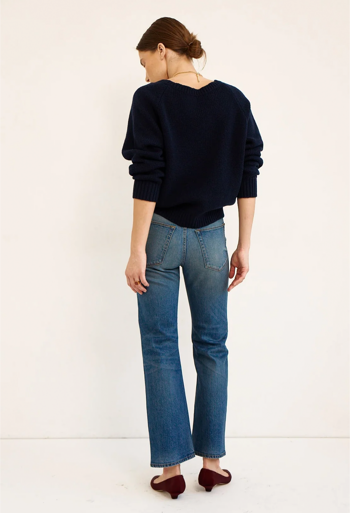 A woman is shown from behind wearing a dark navy blue knit sweater, blue jeans, and burgundy flats against a plain white studio background.