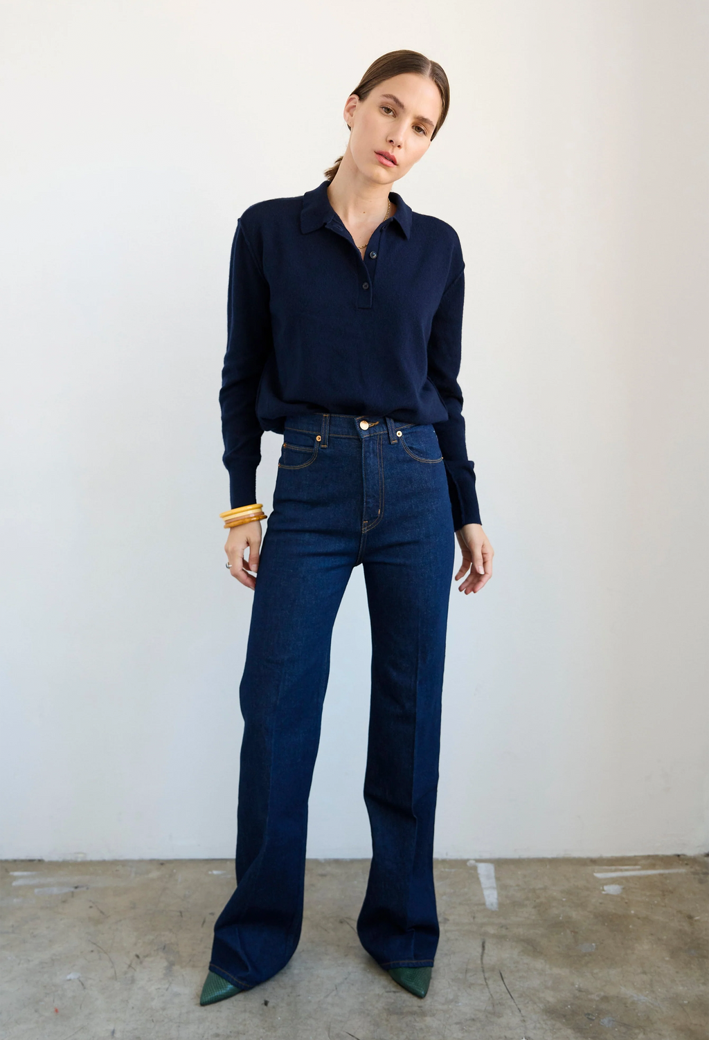 A woman modeling a dark navy long-sleeve knit polo shirt tucked into dark blue high-waisted flare jeans, standing against a plain off-white wall.