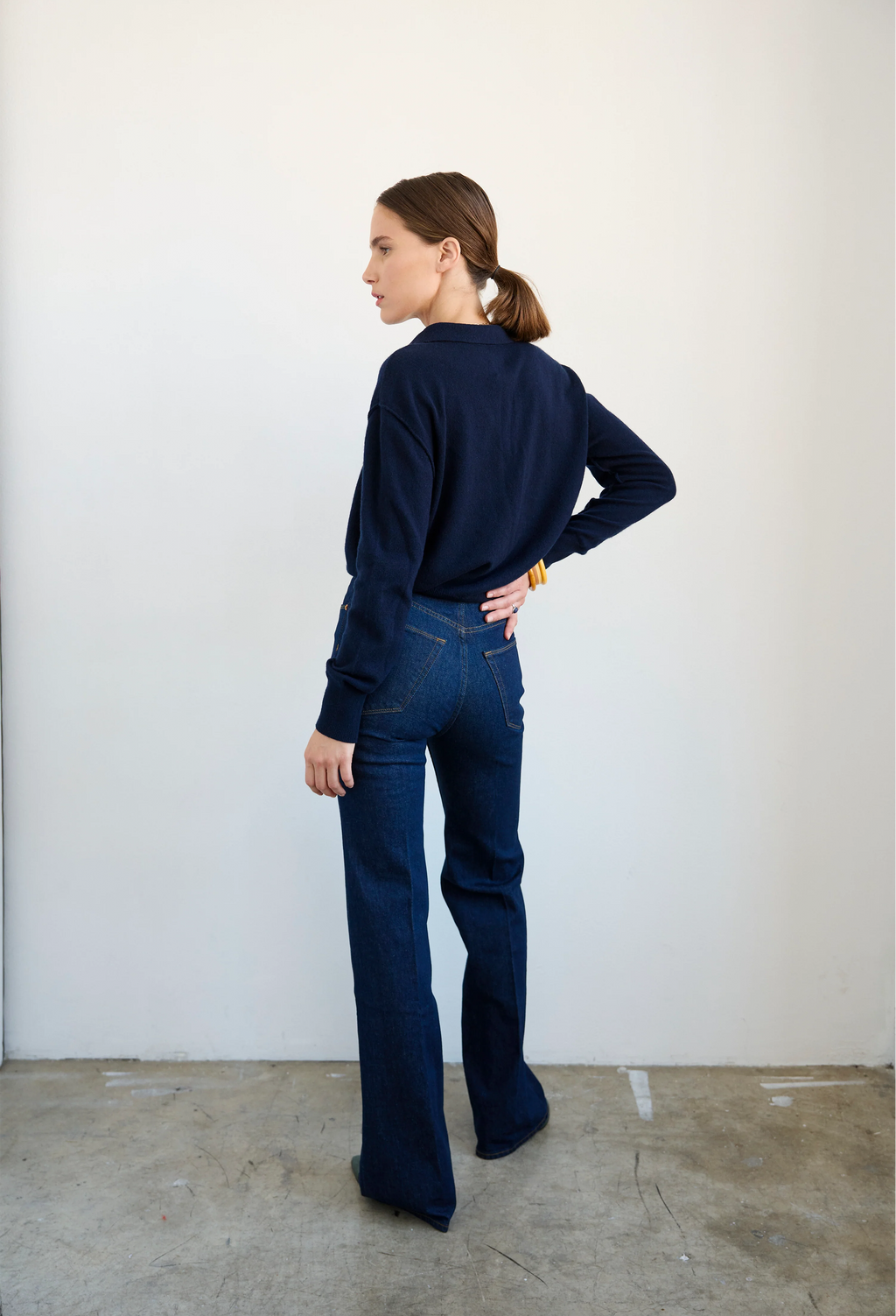 A woman stands in profile wearing a dark navy long-sleeve top and dark blue flared jeans against a plain off-white wall and concrete floor.