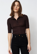 A woman wearing a dark brown ribbed short-sleeve polo shirt with black jeans, standing against a light gray studio background.