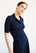 A woman wearing a dark navy ribbed short-sleeve polo shirt with a matching pleated skirt, photographed against a clean white studio background.