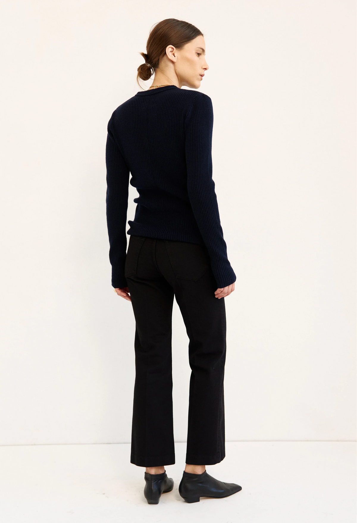 A woman is shown from the back wearing a dark navy ribbed knit sweater, black cropped trousers, and black ankle boots against a plain white studio background.