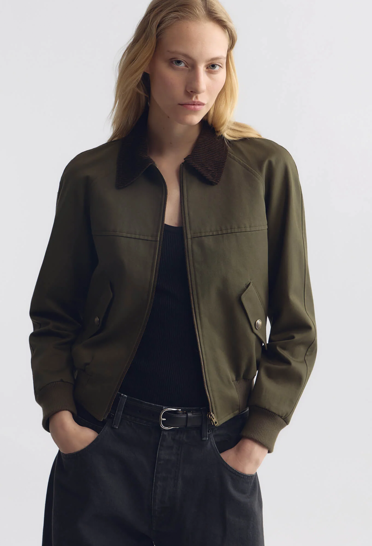 A woman wearing an olive green utility jacket with a brown corduroy collar, styled over a black top and dark jeans against a light gray studio background.