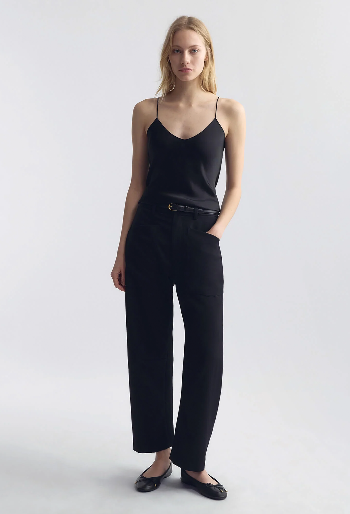 A woman modeling a black camisole top and black wide-leg trousers with a thin black belt, paired with black flats, against a light gray studio background.