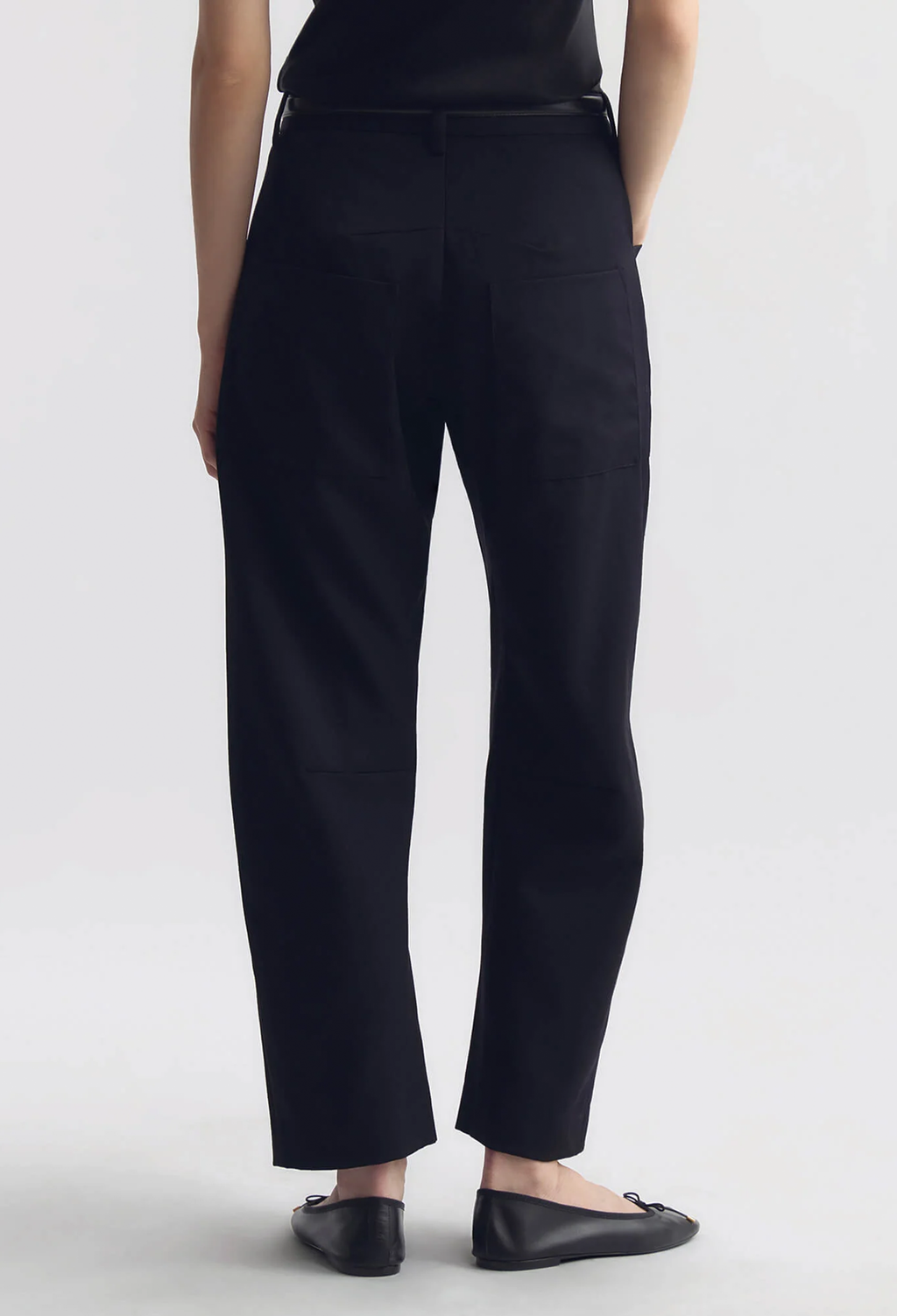A person shown from the back wearing black cropped trousers, a black top, and black flats against a light gray studio background.