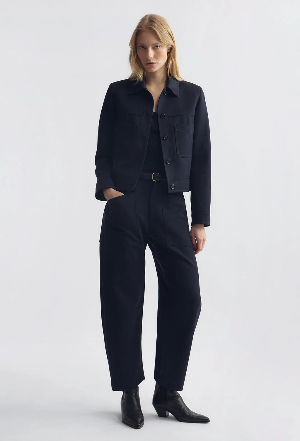 A model stands against a light gray studio background wearing a dark navy blue cropped jacket and matching wide-leg trousers, styled with black ankle boots.