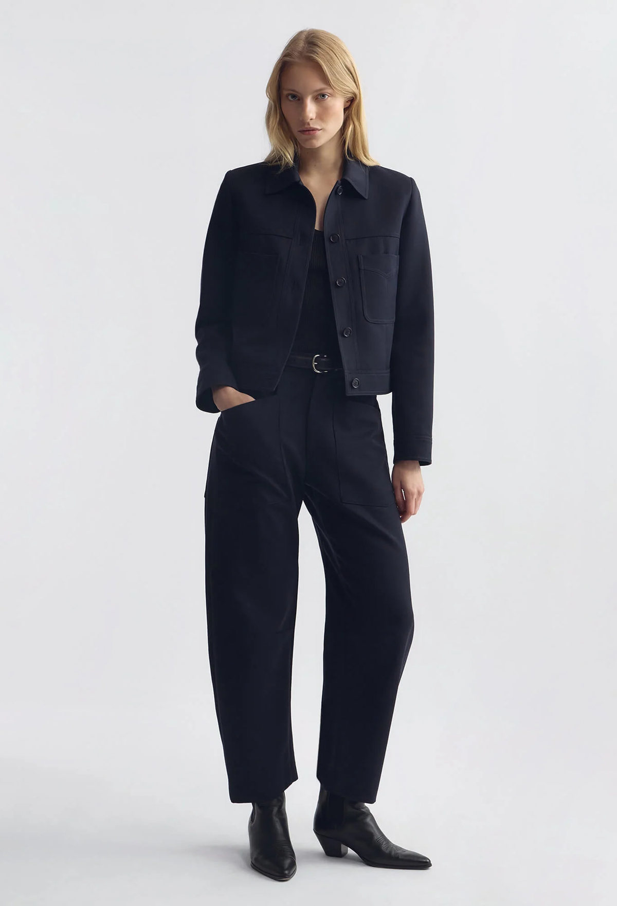 A model stands against a light gray studio background wearing a dark navy blue cropped jacket and matching wide-leg trousers, styled with black ankle boots.