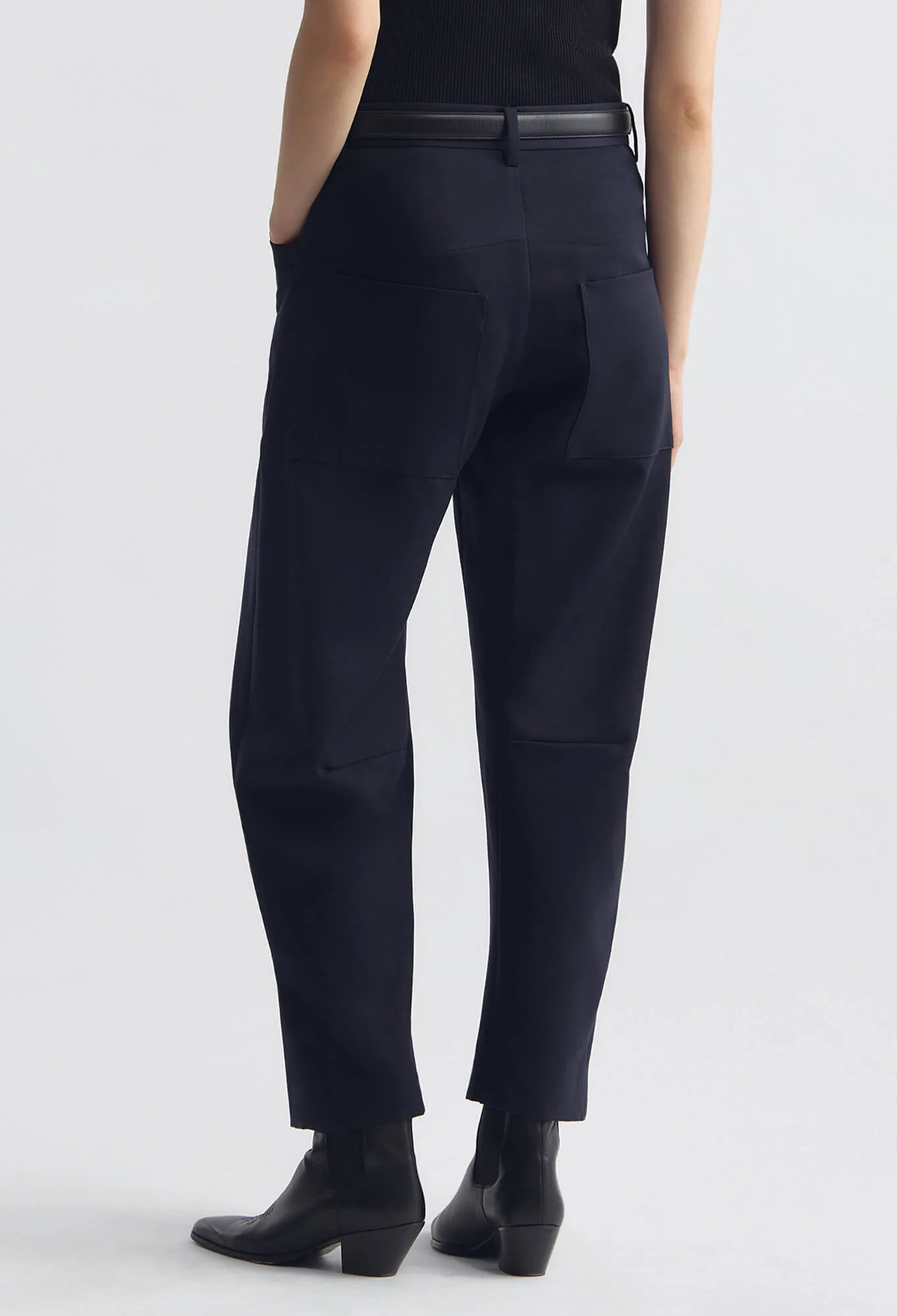 A person shown from the back wearing dark navy tailored trousers with a black belt and black ankle boots against a light gray studio background.