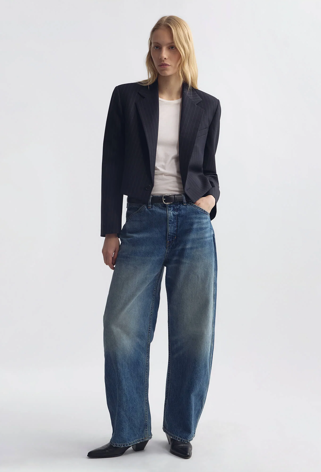 A model stands against a light gray studio background wearing a dark navy blazer, a white top, wide-leg blue jeans, and black shoes.