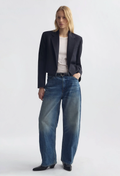 A model stands against a light gray studio background wearing a dark navy blazer, a white top, wide-leg blue jeans, and black shoes.