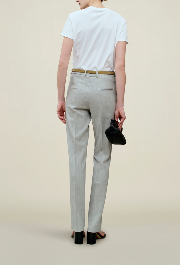The back of a woman wearing a light grey pants, a white top, and a brown belt. Beige background.