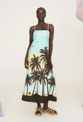 A woman wearing a light blue midi dress with black palm trees and thin straps. White background.
