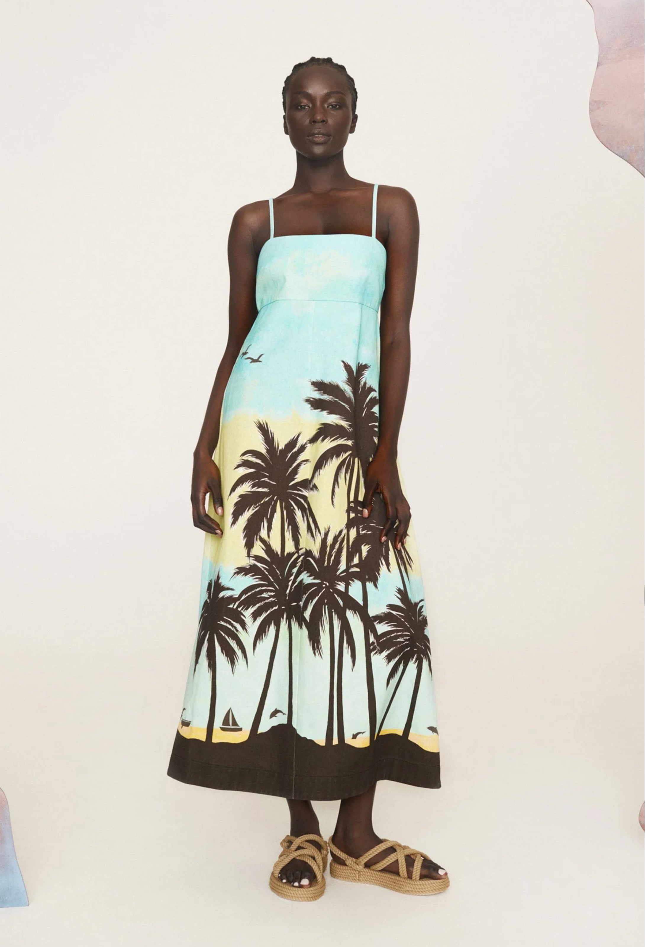 A woman wearing a light blue midi dress with black palm trees and thin straps. White background.