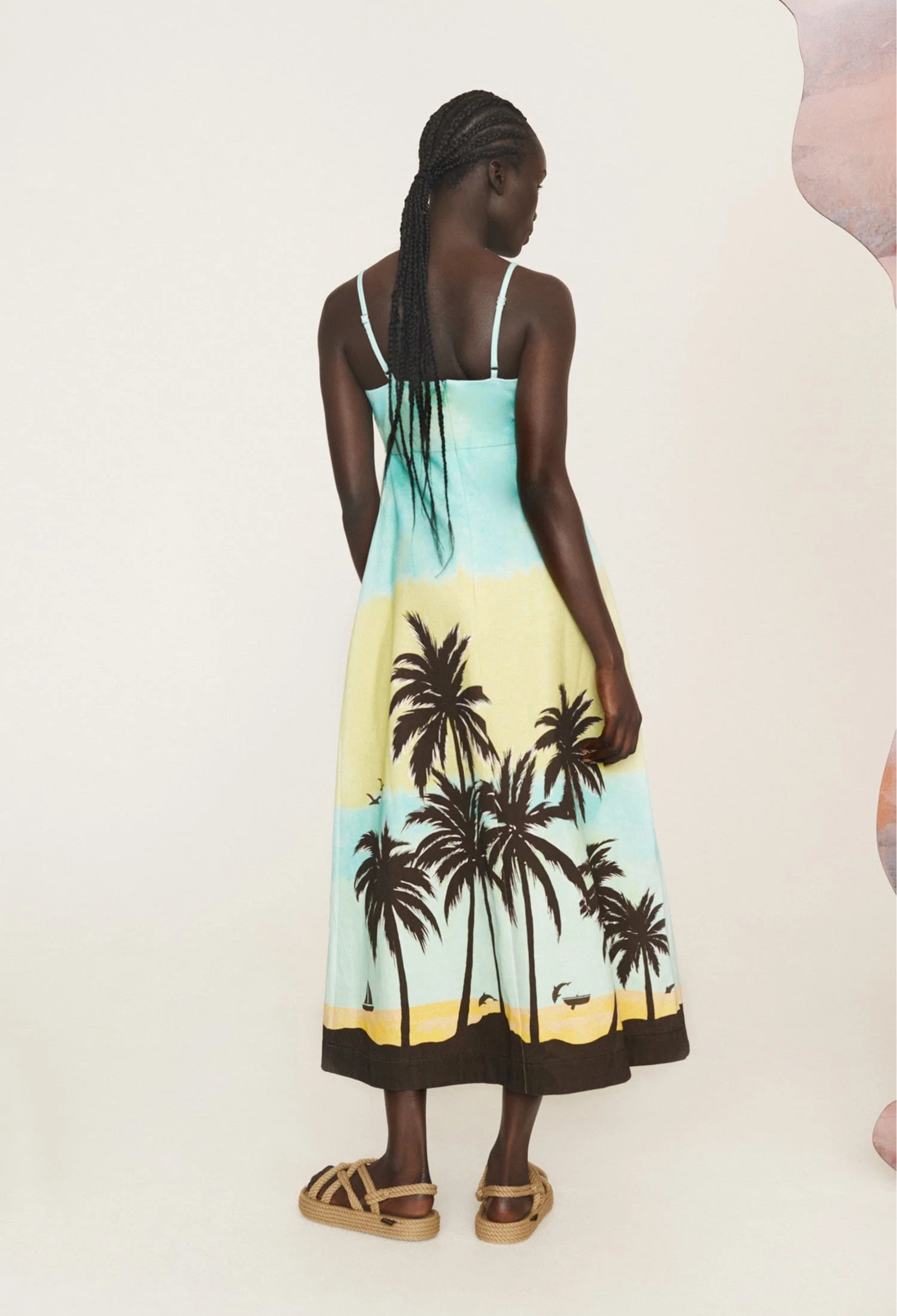 The back of a woman wearing a light blue midi dress with black palm trees and thin straps. White background.