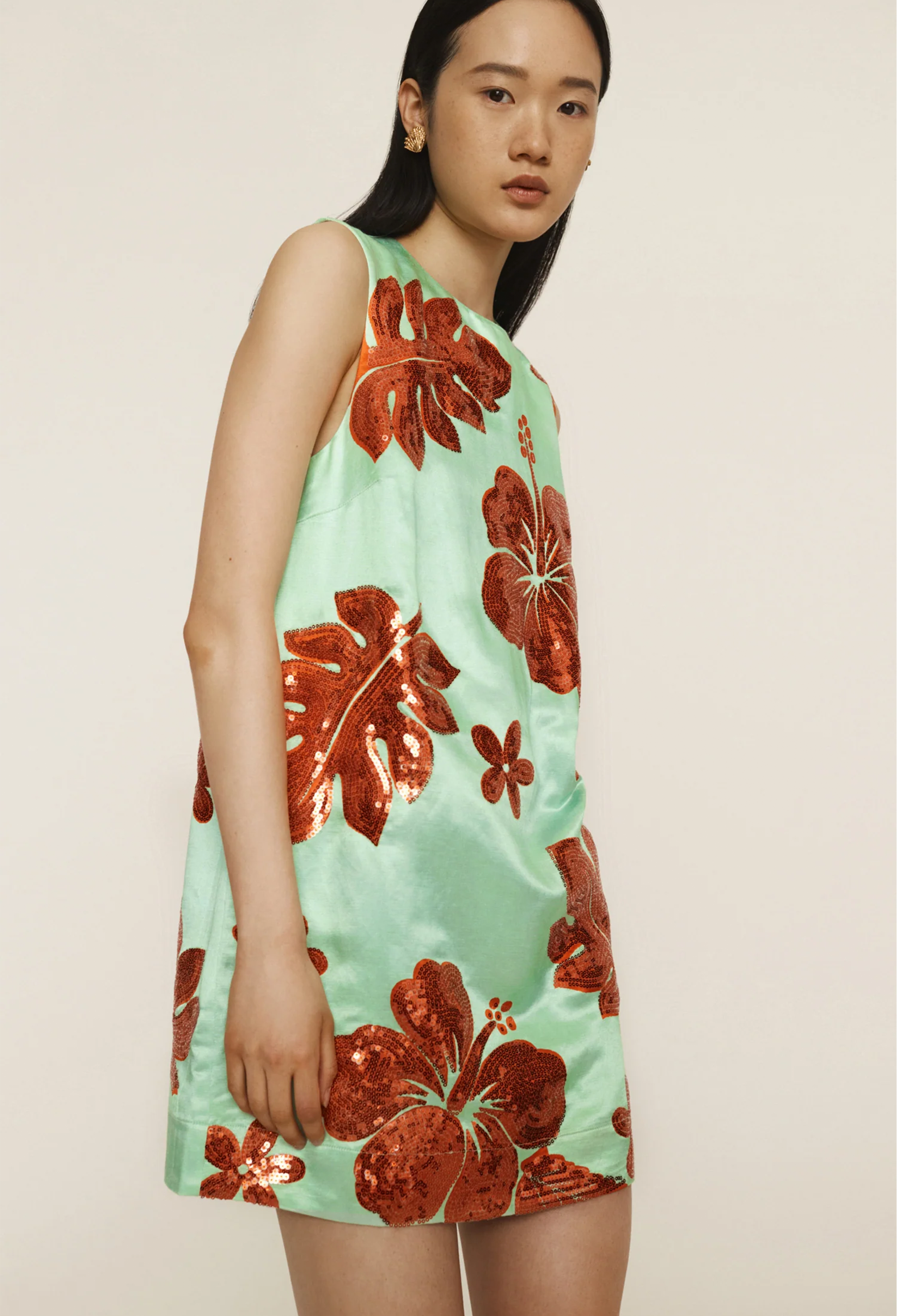 A woman wearing a green mini dress with orange sequined leaves and flowers all over. White background.