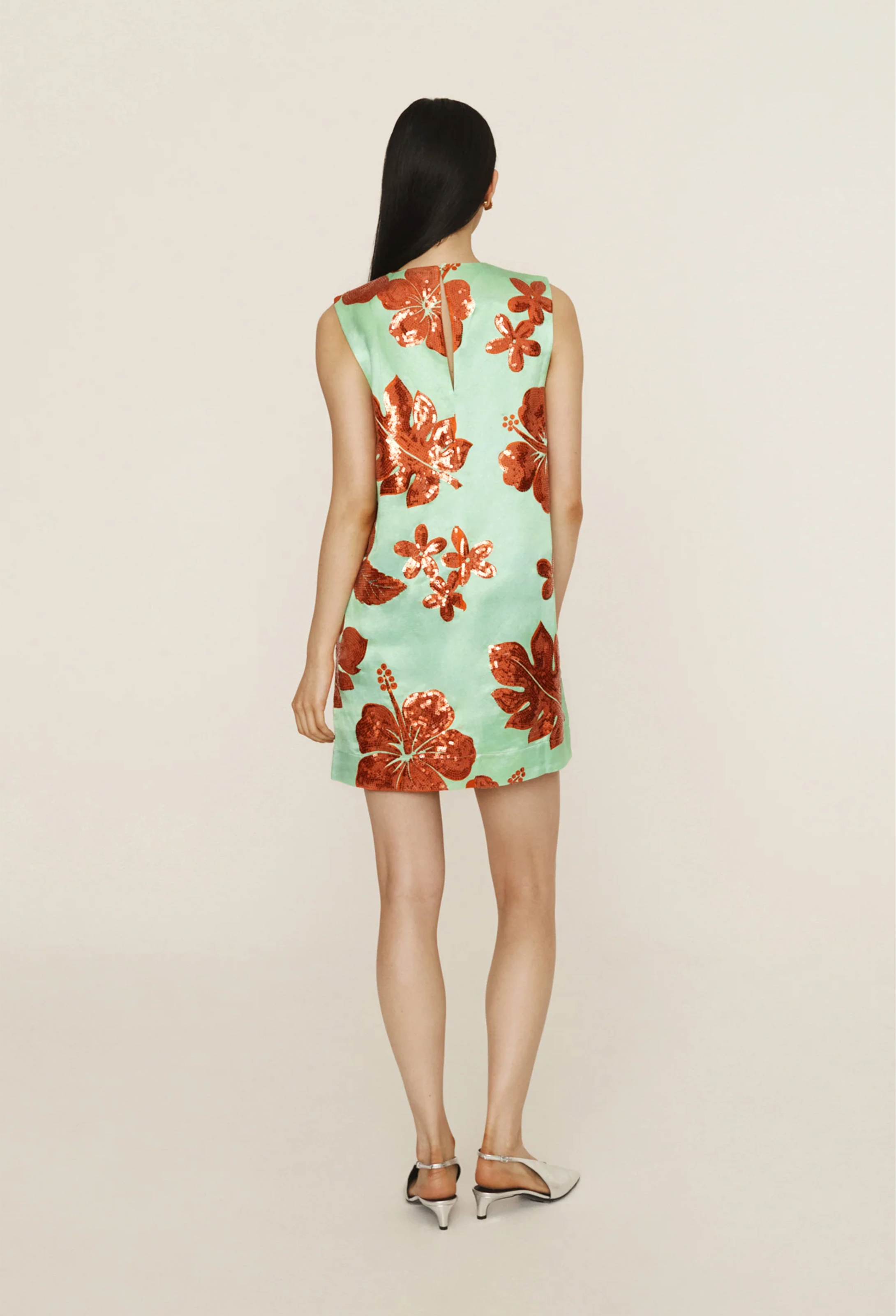 The back of a woman wearing a green mini dress with orange sequined leaves and flowers all over. White background.