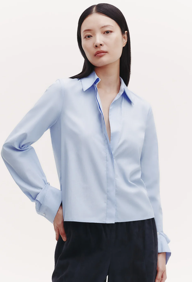 A woman wearing a slightly cropped light blue button down shirt with a covered placket. White background.
