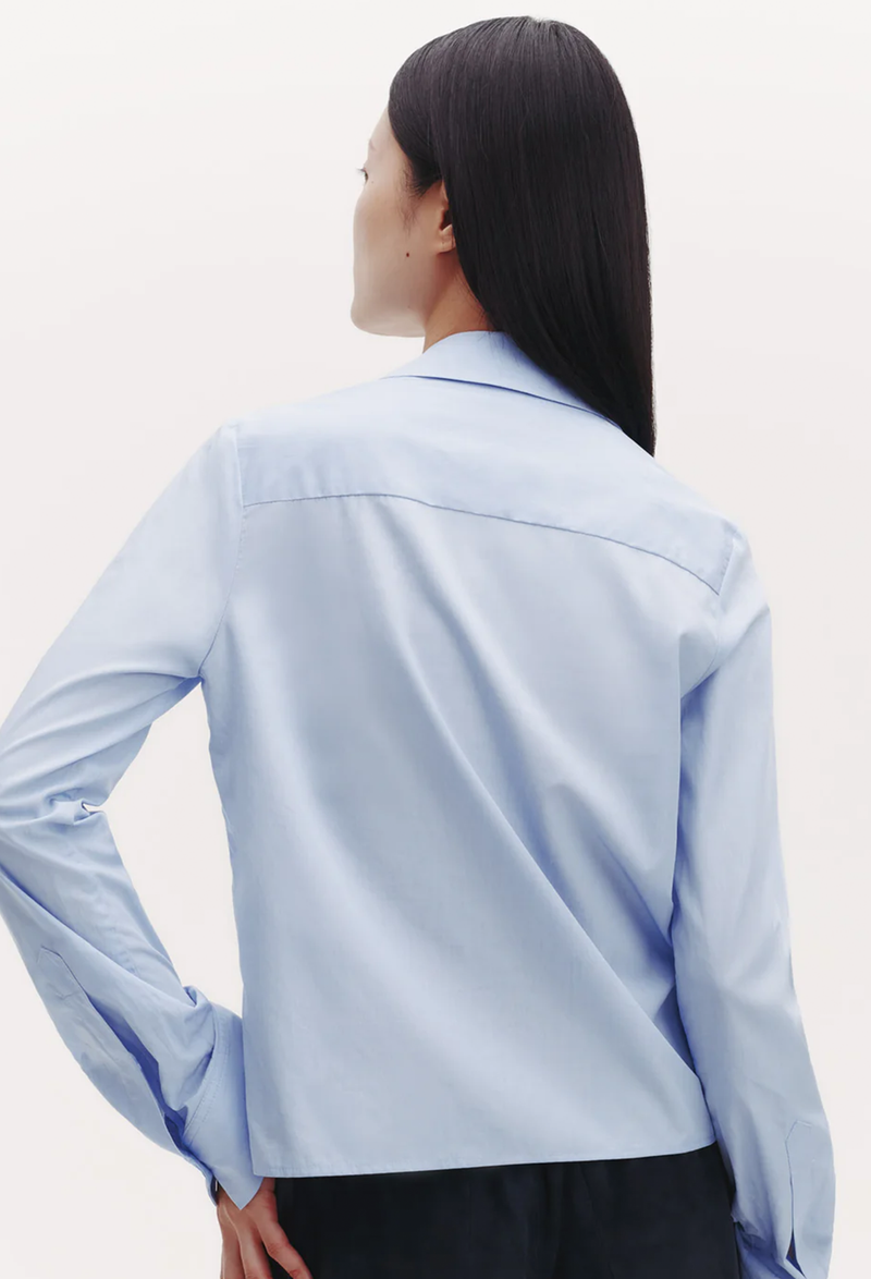 The back of a woman wearing a slightly cropped light blue button down shirt with a covered placket. White background.