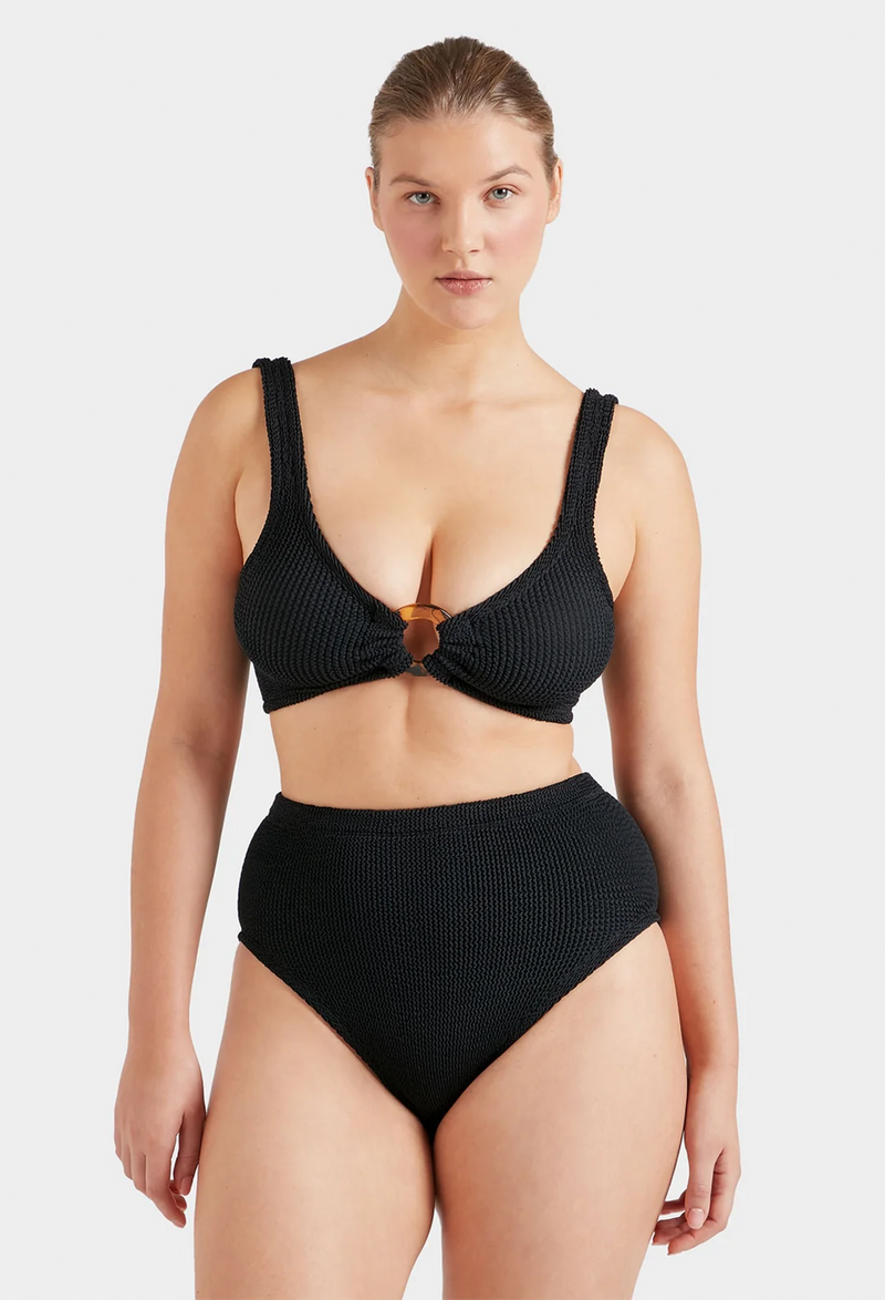 A woman wearing a black two-piece swimsuit with a tortoiseshell hoop gathering the top and high waisted bottoms. White background.