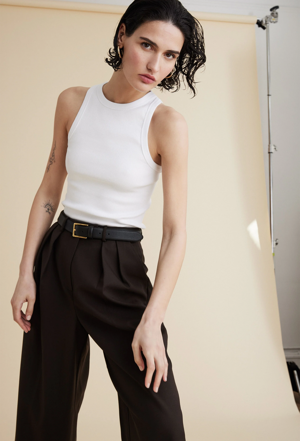 A woman wearing a white ribbed tank top and brown pants. Beige background.