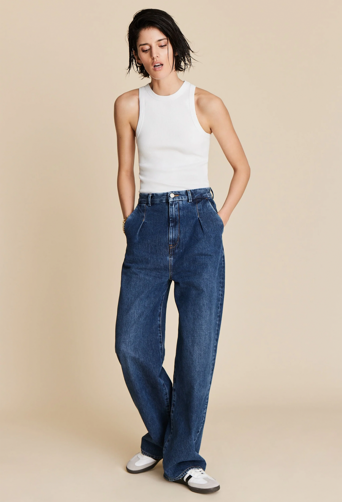 A woman wearing a white ribbed tank top and jeans. Beige background.