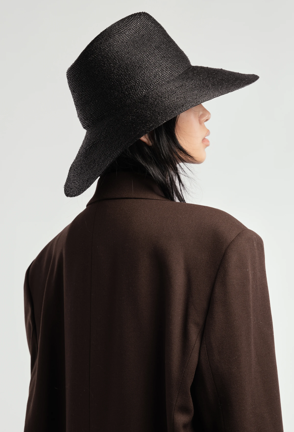 The back of a woman wearing a black straw hat with a downward tilted. brim and brown blazer. White background.