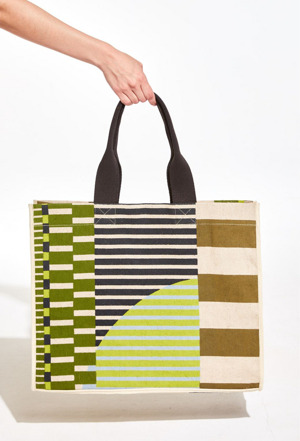 A hand holding a large green, blue, and off-white striped tote bag with black handles. White background.