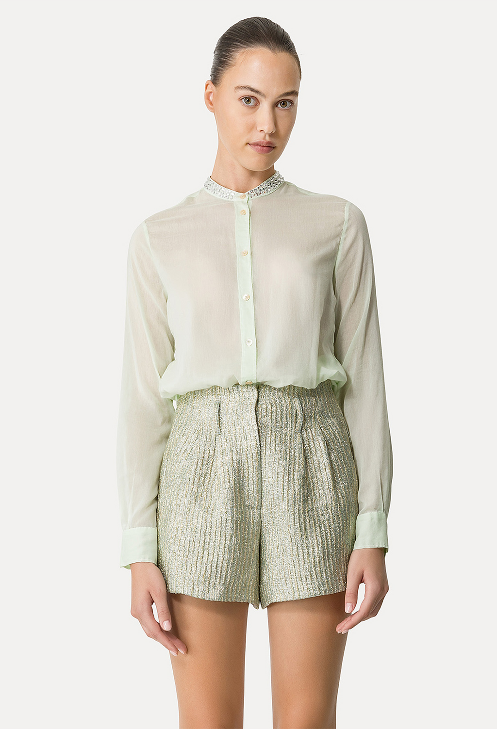 A woman wearing a green long sleeve button down sheer shirt tucked into matching metallic shorts. White background.