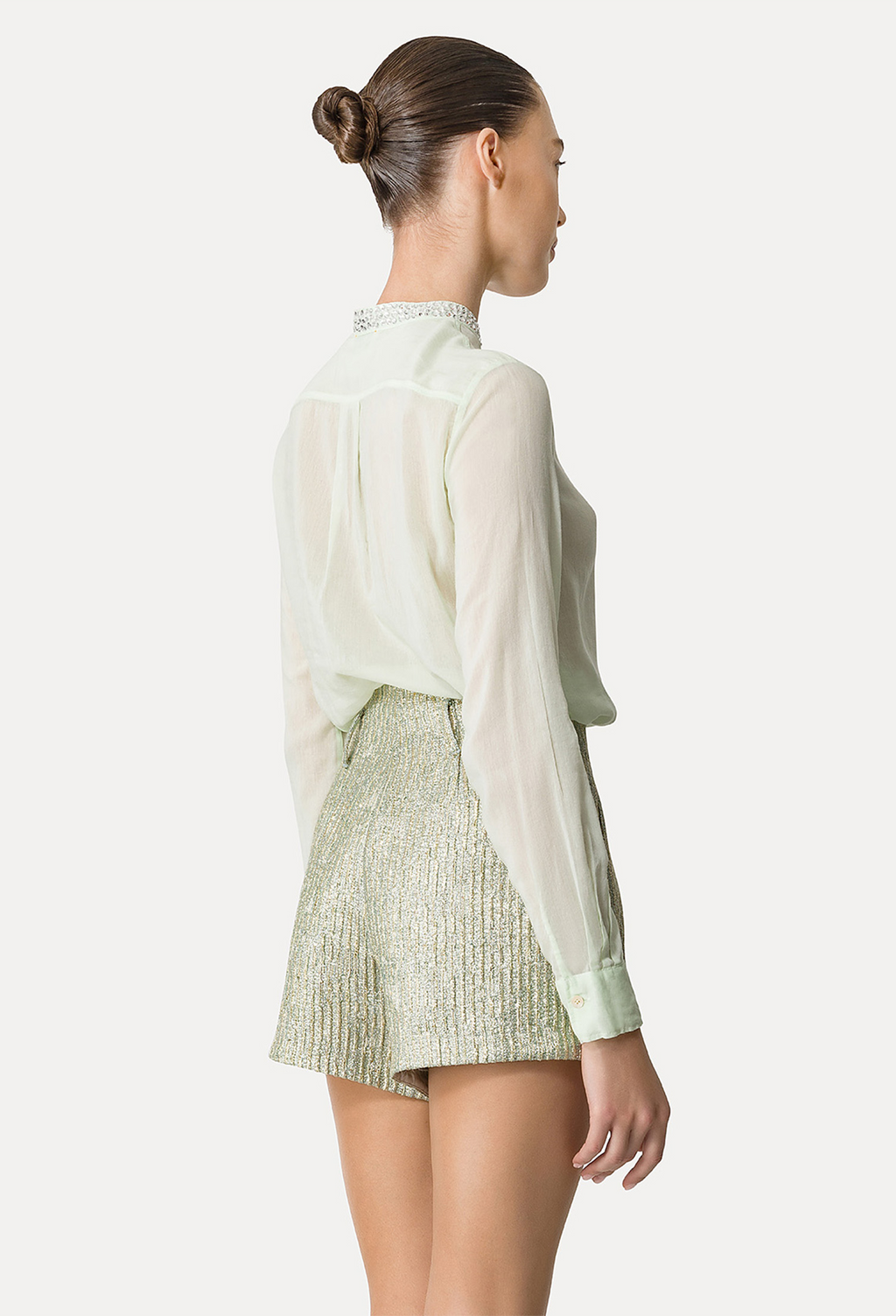 The back of a woman wearing a green long sleeve button down sheer shirt tucked into matching metallic shorts. White background.