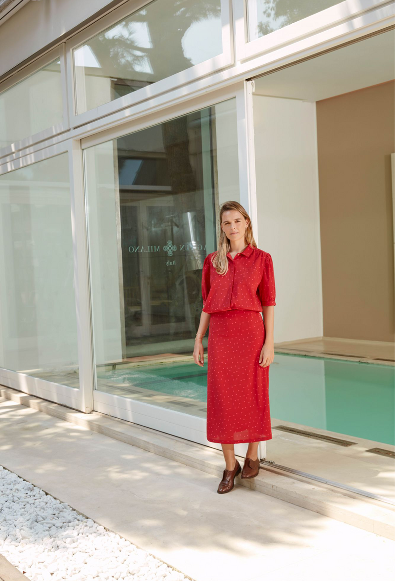 A woman outside of a store wearing a half sleeve red button down shirt with pink polka dots and a shirt pocket and matching long skirt.