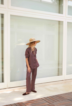 A woman in a hat outside of a building with a matching brown button down with white polka dots and pants.