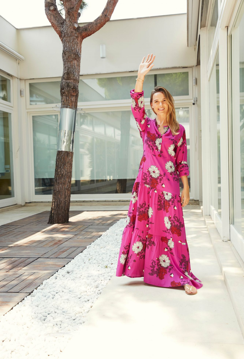 A woman outside of a building in front of a tree waving while wearing a long floral hot pink dress with a tiered skirt and long sleeves.
