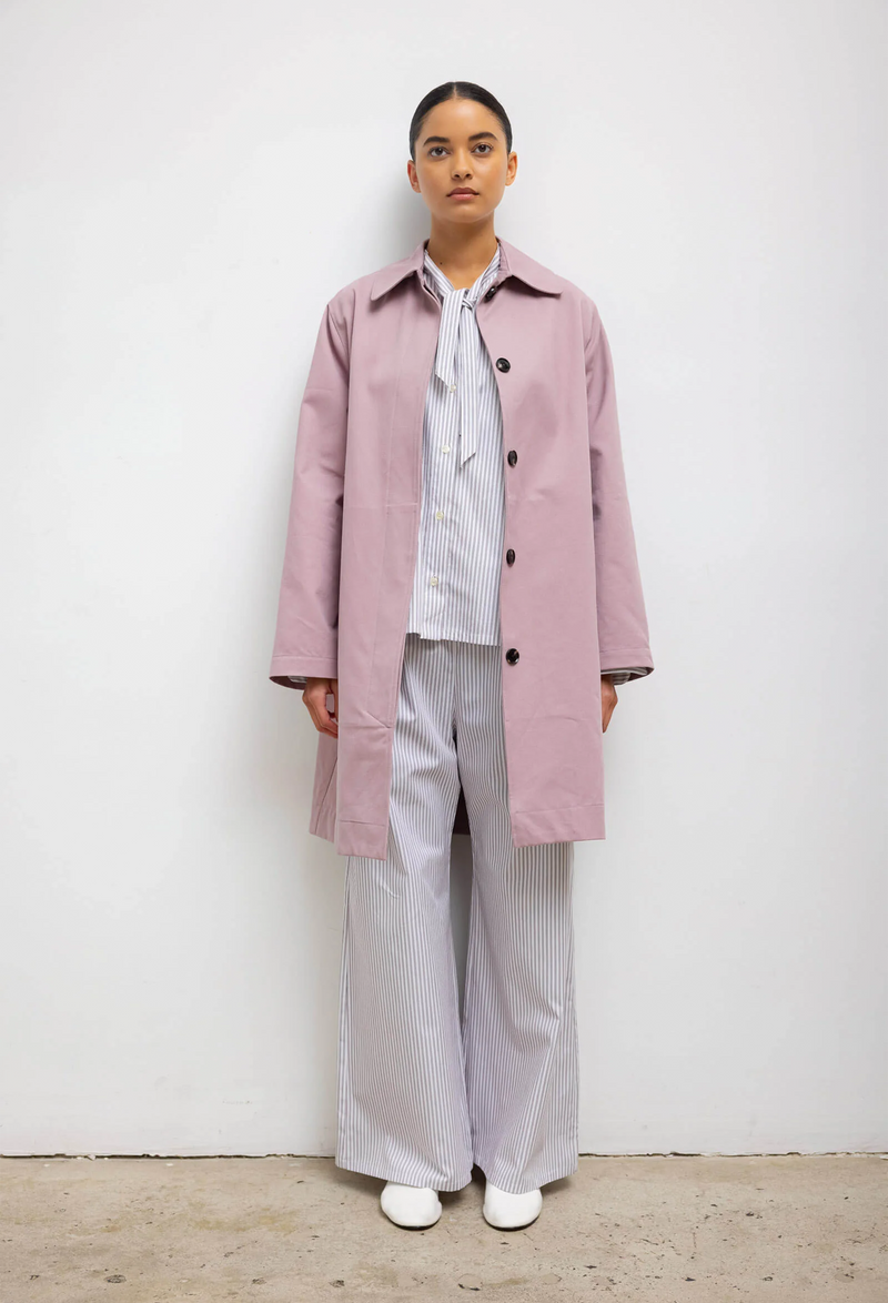 A woman wearing a blue and white striped shirt and pants set with a pink overcoat. White background.