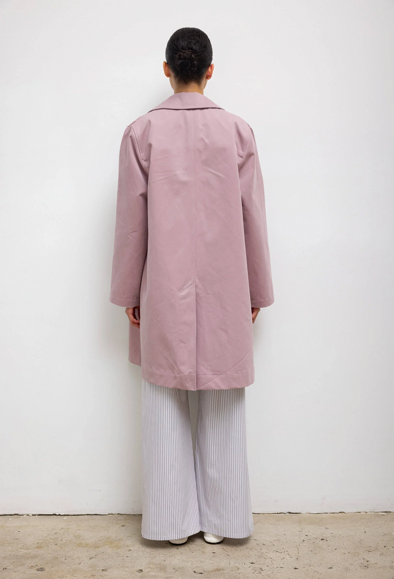 The back of a woman wearing a blue and white striped shirt and pants set with a pink overcoat. White background.