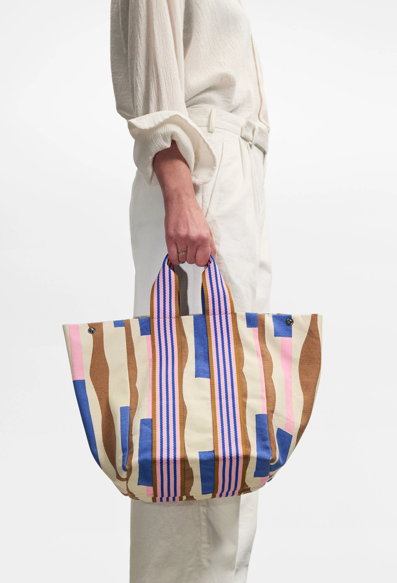 A person holding a canvas tote bag with a brown, pink, and blue print on it. White background.