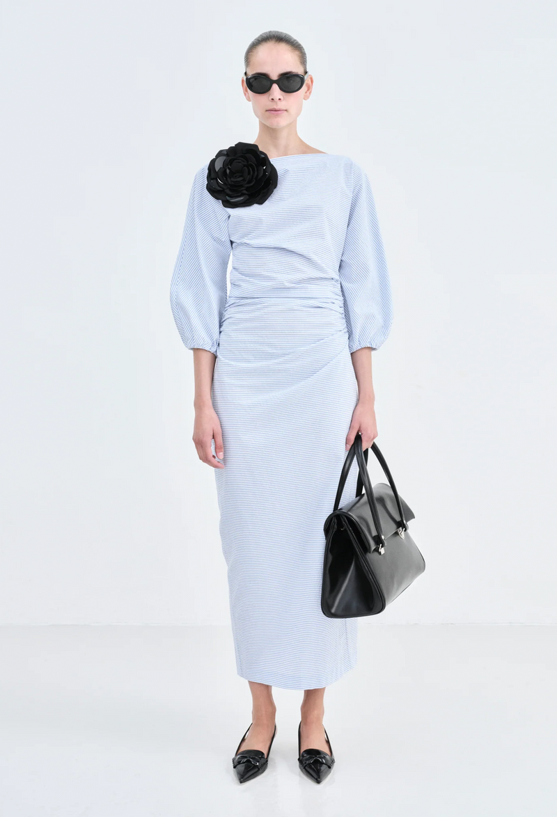 A woman with a black bag wearing a long sleeved cinched waist long dress with blue and white stripes and a large black flower brooch near her shoulder. White background.