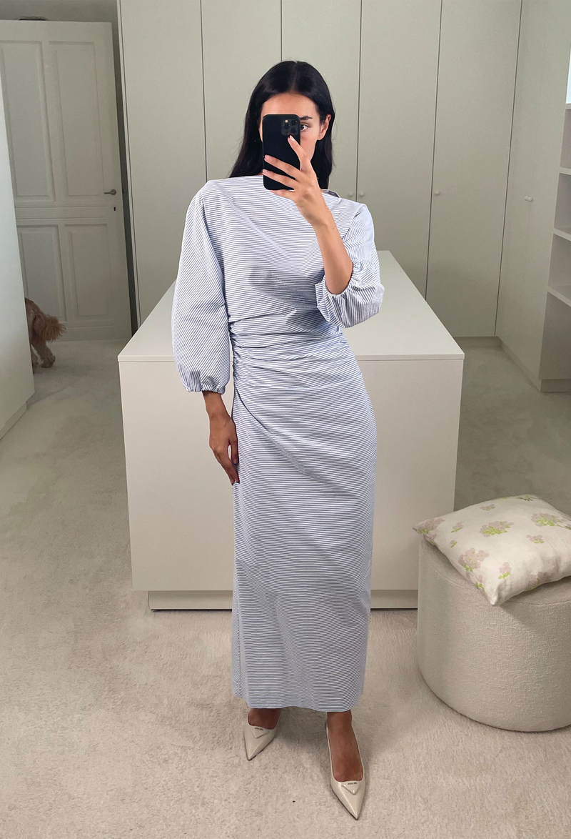 A woman in a closet taking a mirror selfie wearing a long sleeved cinched waist long dress with blue and white stripes.