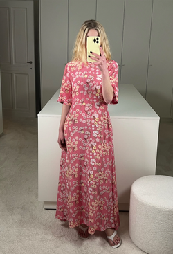 A woman in a closet taking a mirror selfie wearing a short sleeve long pink-red dress with a pastel colored flowers print.