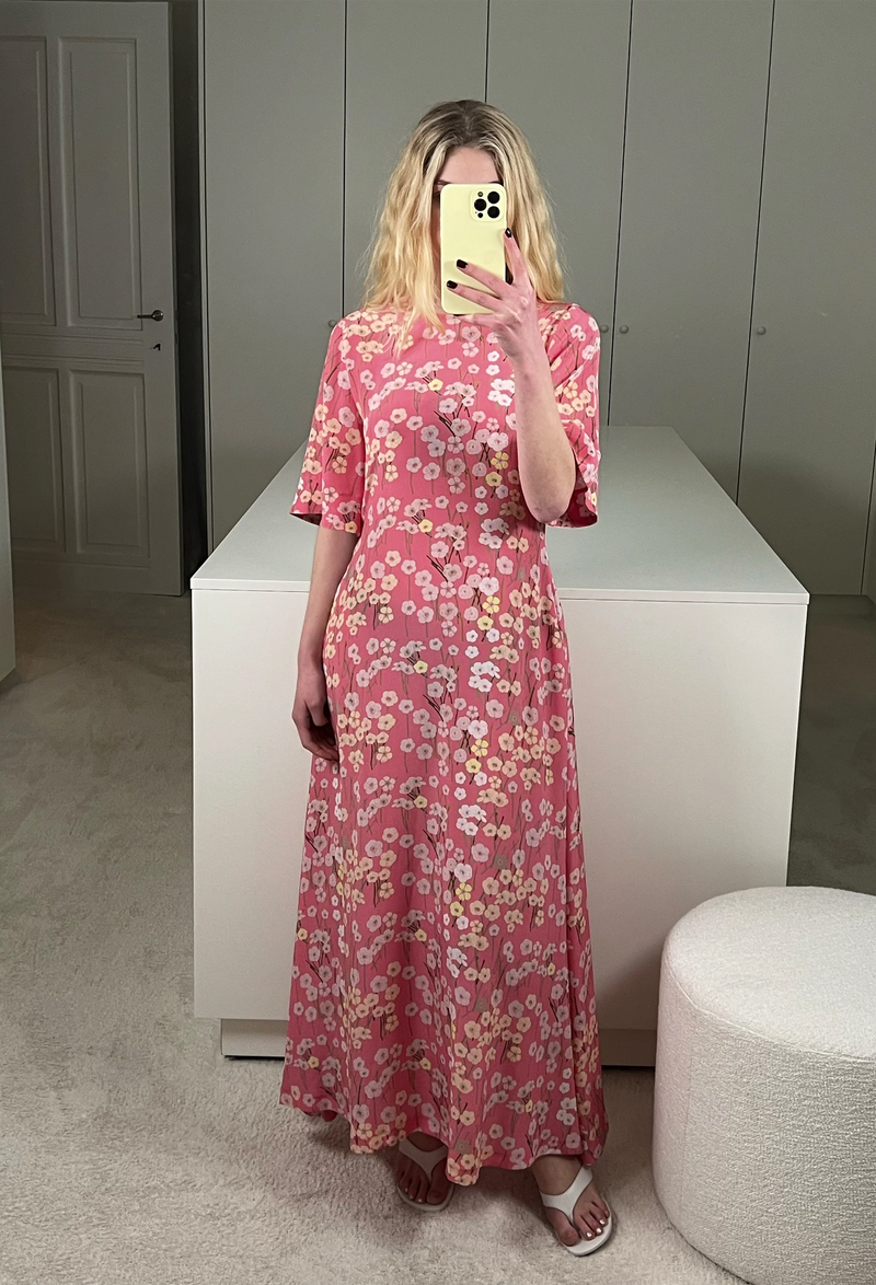 A woman in a closet taking a mirror selfie wearing a short sleeve long pink-red dress with a pastel colored flowers print.