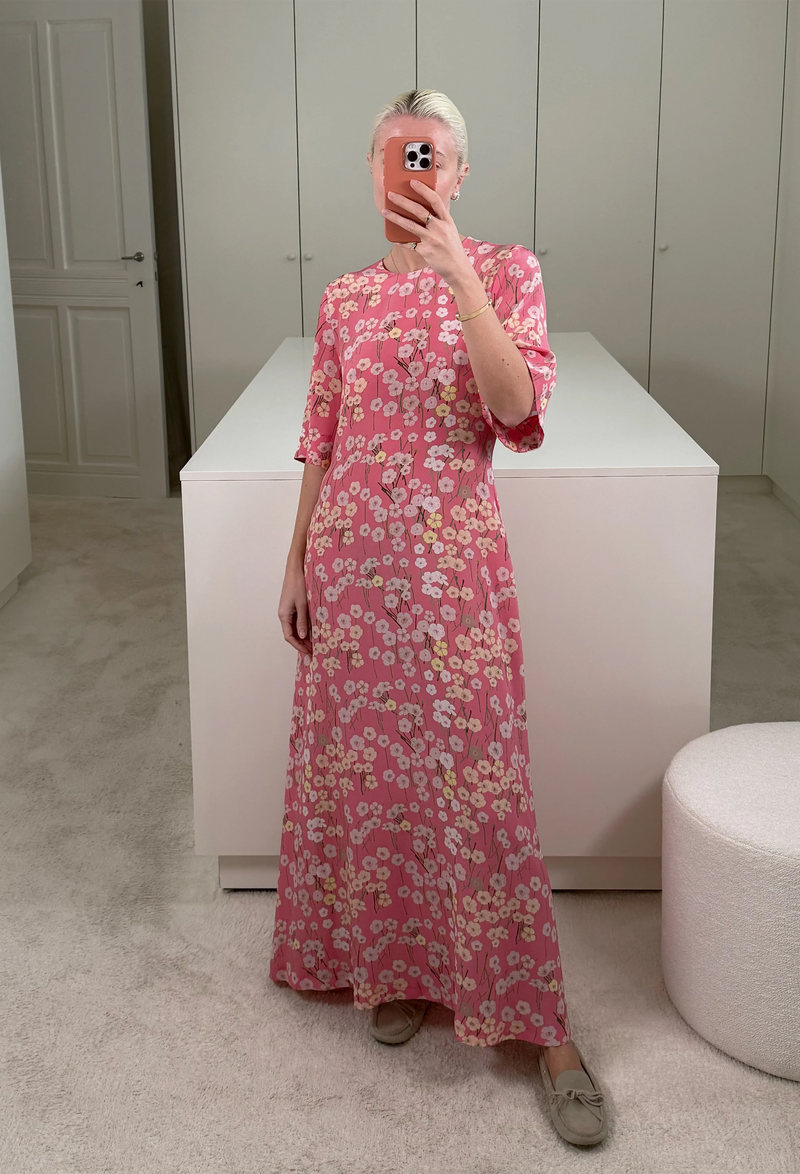 A woman in a closet taking a mirror selfie wearing a short sleeve long pink-red dress with a pastel colored flowers print.