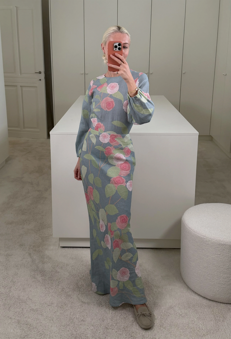 A woman in a closet taking a mirror selfie wearing a blue dress with long sleeves and pink and red roses on it.
