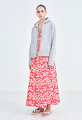 A woman wearing a grey zip-up jacket and a long red dress with a pastel colored flowers print. White background.