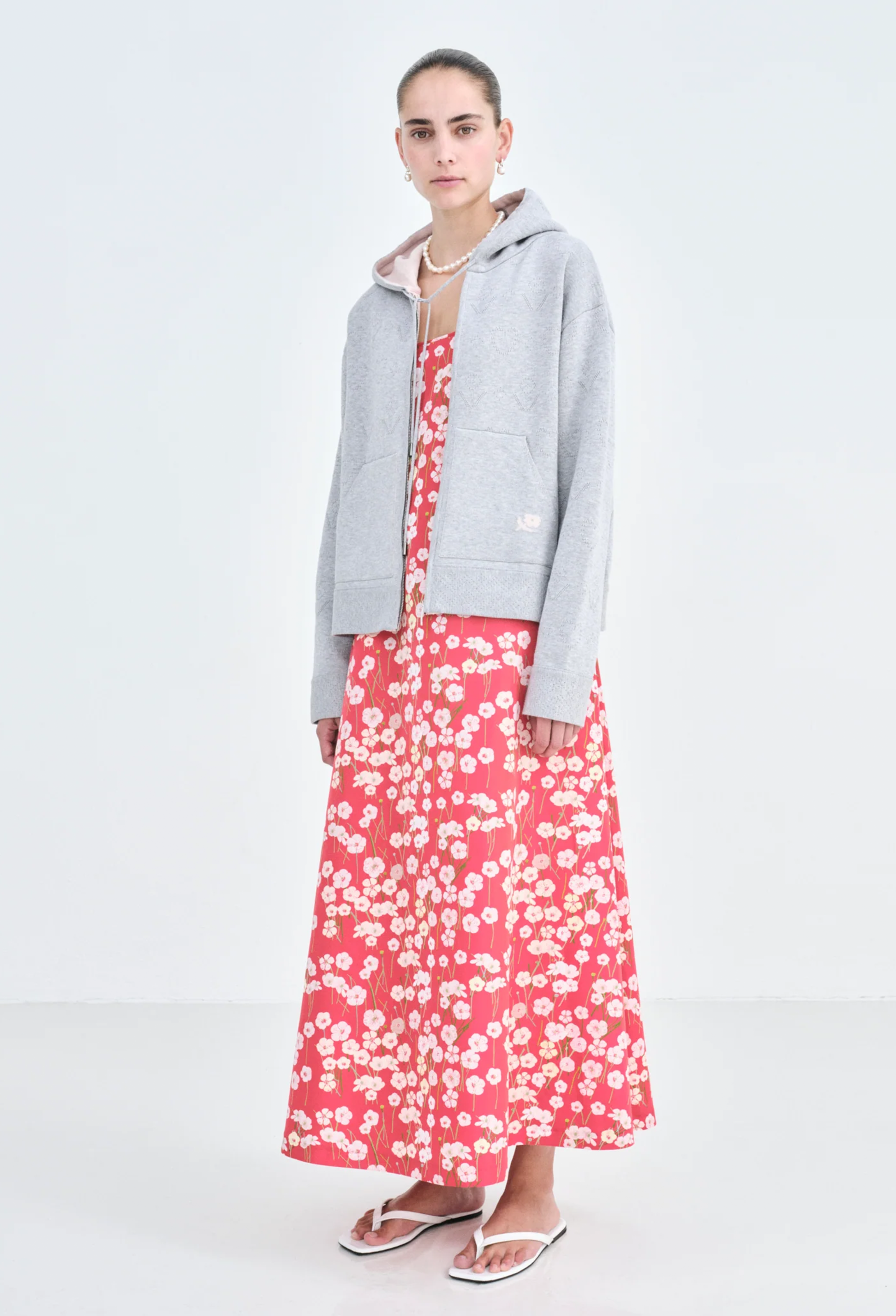 A woman wearing a grey zip-up jacket and a long red dress with a pastel colored flowers print. White background.