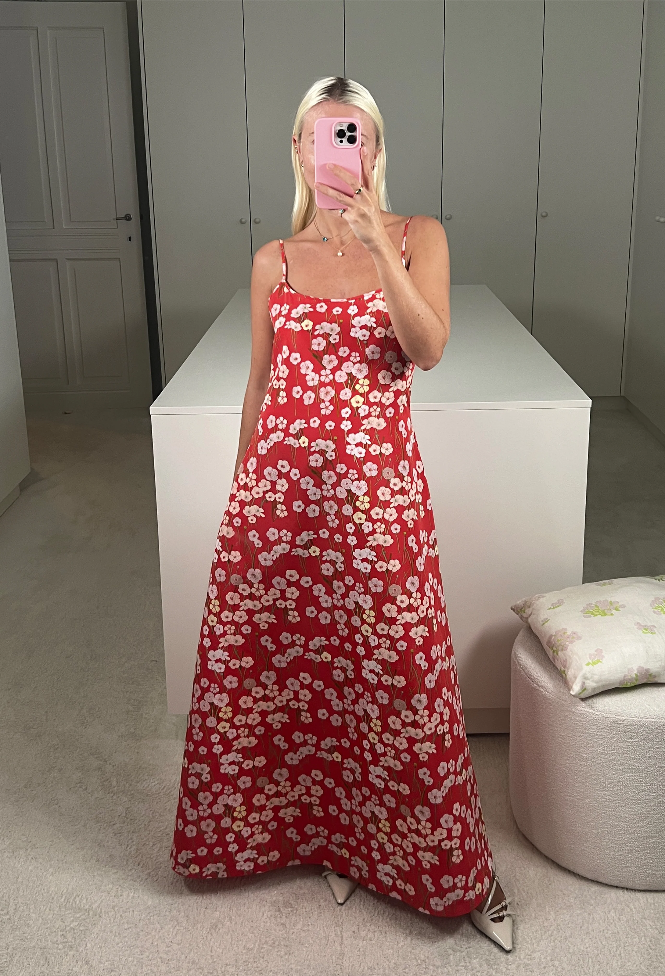 A woman in a closet taking a mirror selfie wearing a red dress with a pastel colored flowers print and spaghetti straps.